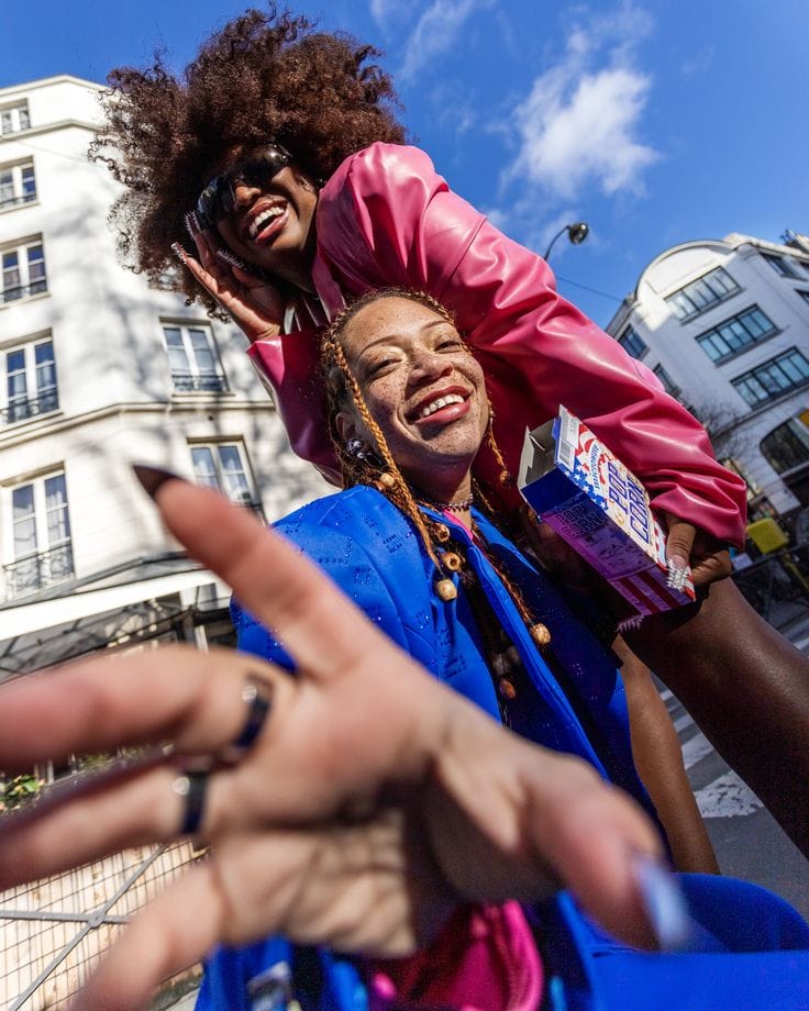 Two friends posing on a city street, one reaching toward the camera