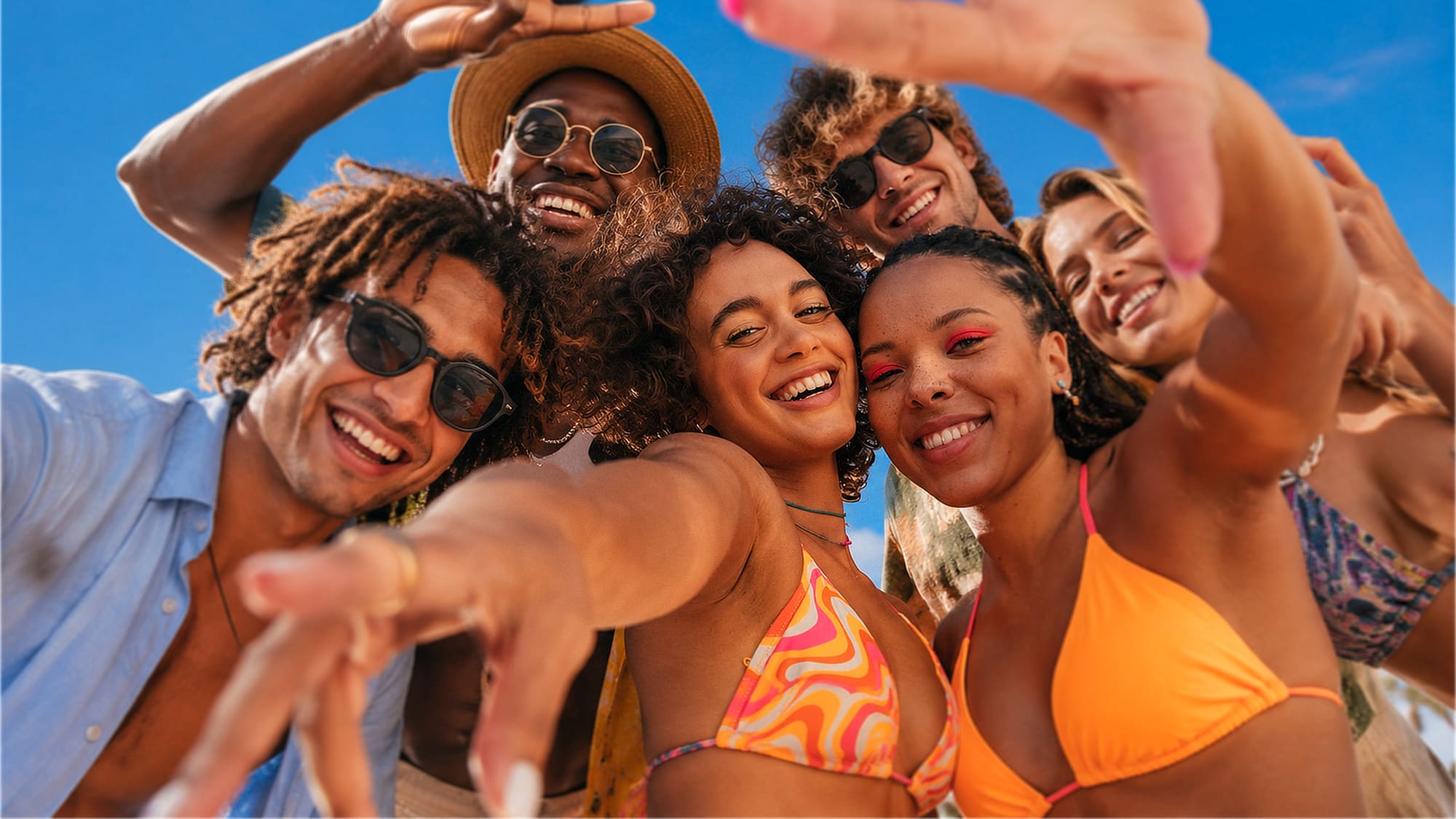 A group of friends leaning into the camera together, arms around each other, shot from below against a bright sky